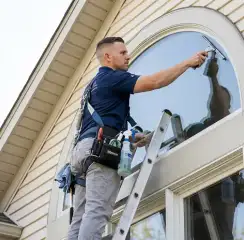 Professional window cleaner on a ladder using a squeegee to clean an exterior arched window, Charleston, SC.